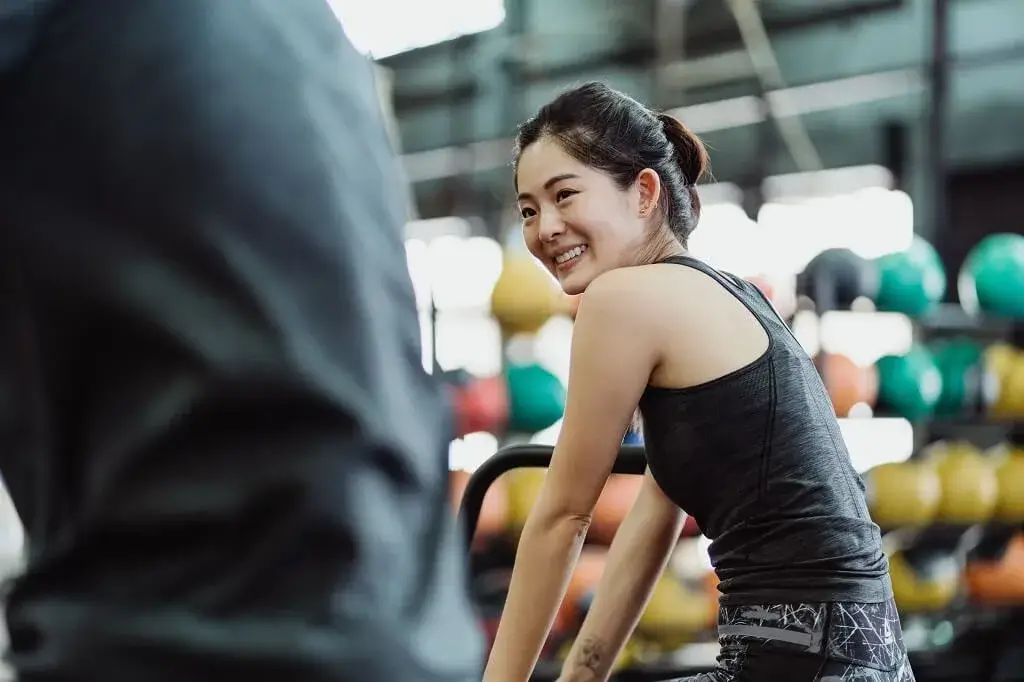 Smiling Asian woman in a gym talking with her coach during a personal training package consultation session.