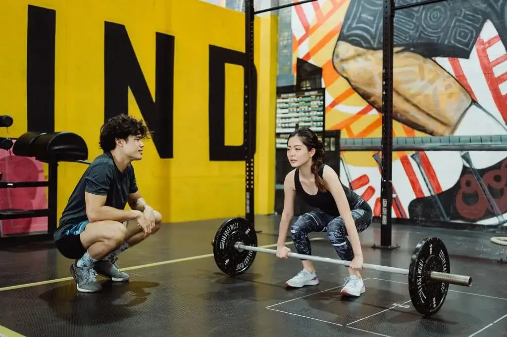 Female client lifting a barbell while coach supervises during a fitness training and strength workout session.