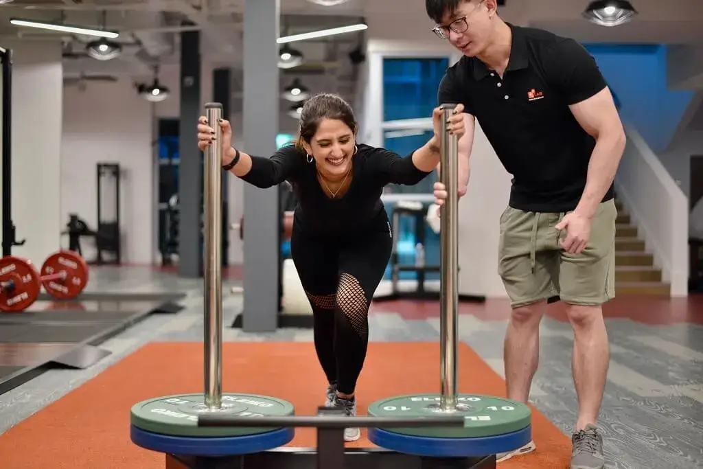 Asian male fitness coach guiding a female client during a private personal training workout session in the gym.