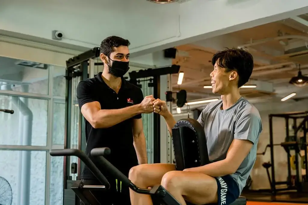 Happy young Asian man laughing and fist bumping his personal trainer after a successful workout session at the gym.